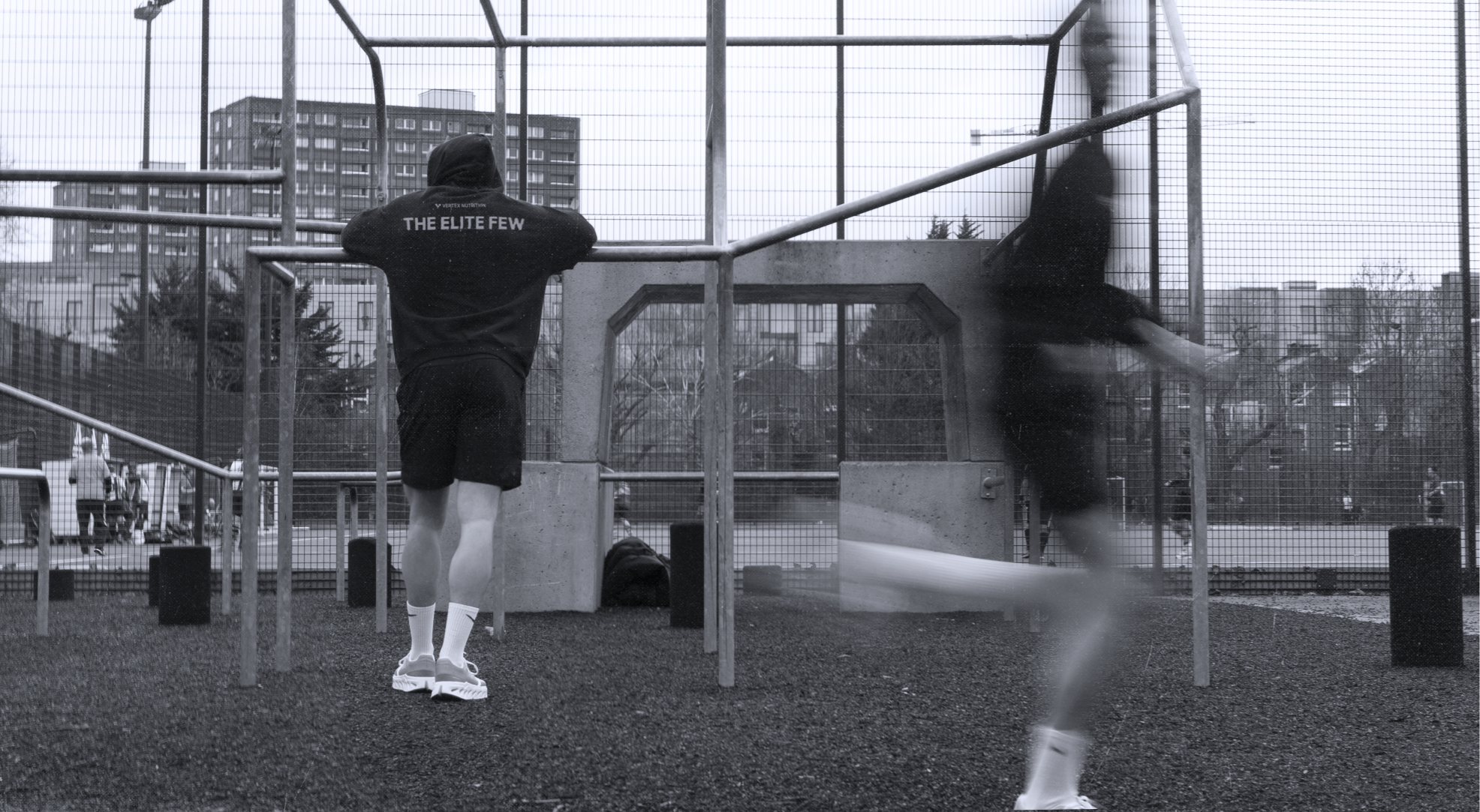 Two individuals exercising outdoors on a fenced court. One person leans on bars with their back facing the camera, while another is blurred in motion running.