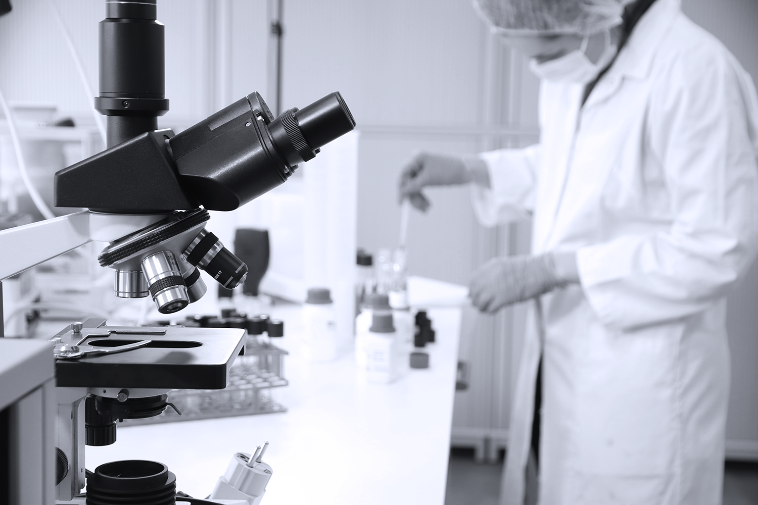 A close-up of a microscope in a laboratory setting, with a researcher in the background handling samples, wearing a lab coat, mask, and gloves.